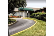 Exterior view of a single-story nursing home building with a curved driveway leading to the entrance, which is covered by a green awning. There are bushes and trees surrounding the building and a well-maintained lawn.