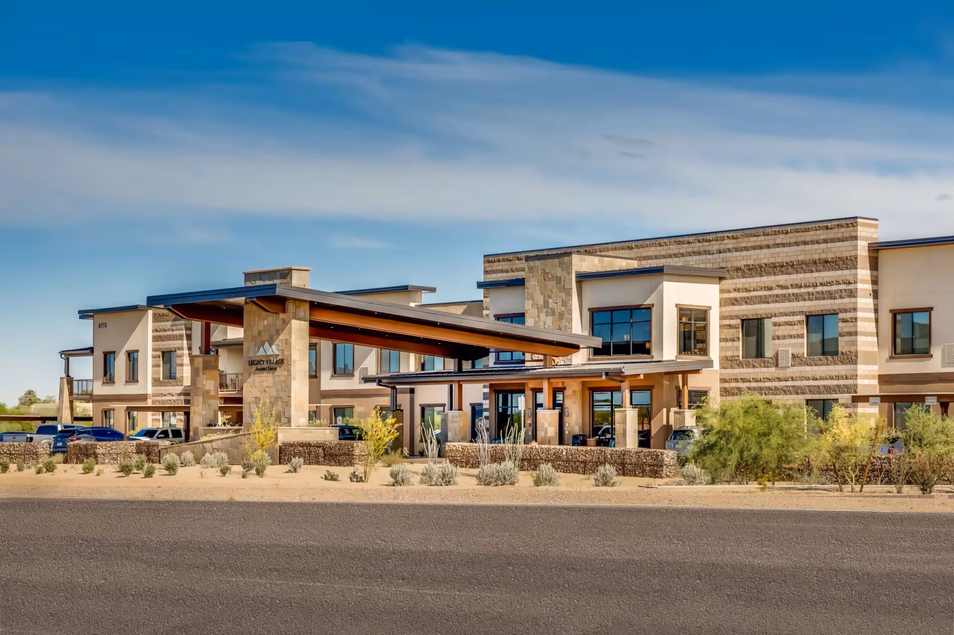 Front exterior of a modern two-story senior living building with a covered porte-cochere, desert landscaping, and parked cars.