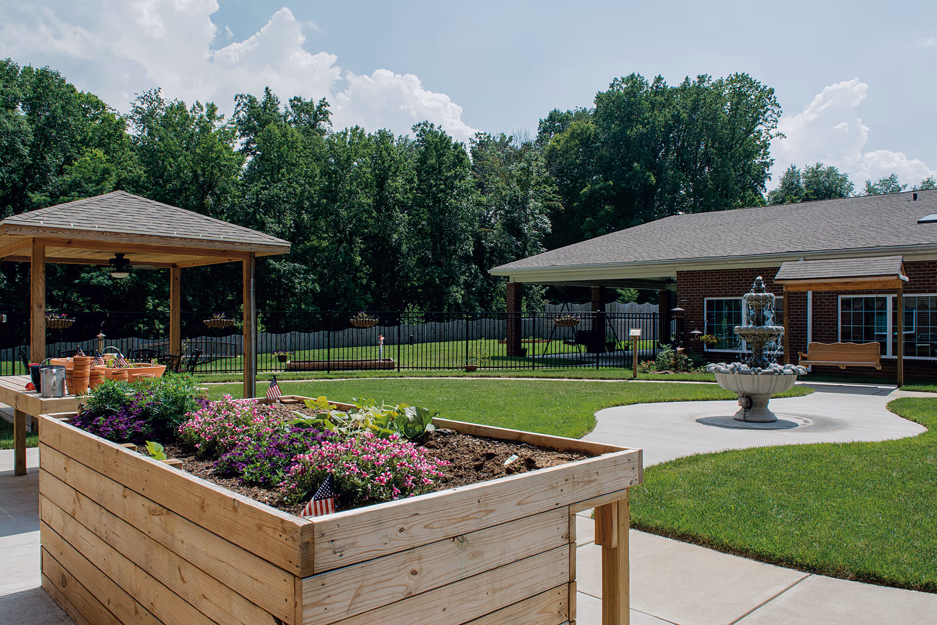 A senior living courtyard with a raised wooden planter filled with flowers, a gazebo, a fountain, and a brick building in the background.