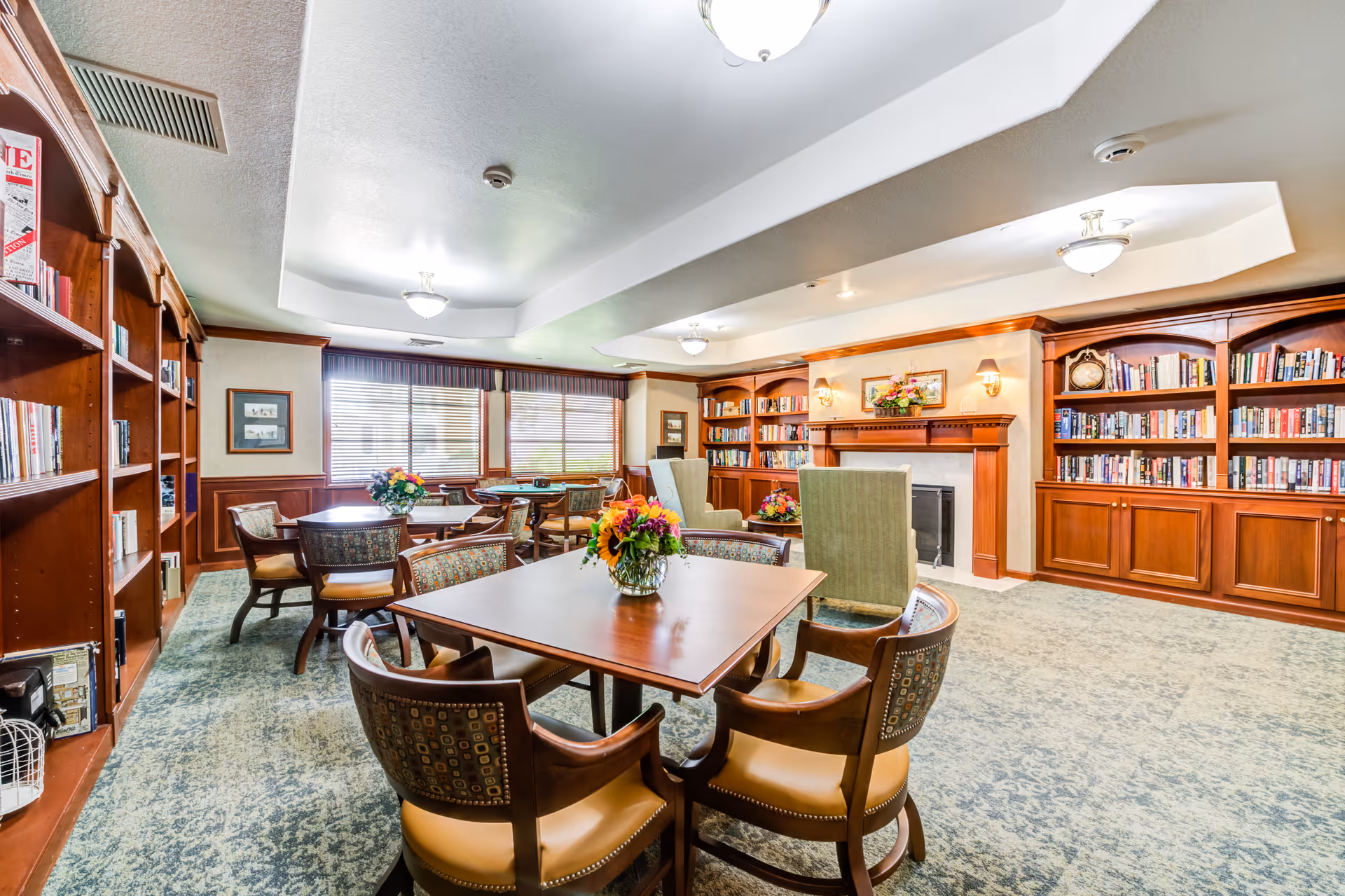 A cozy and well-lit common room with wooden bookshelves filled with books, several tables with chairs arranged around them, and floral centerpieces on the tables. The room features a fireplace with a decorative mantle, two green upholstered armchairs, and large windows with blinds allowing natural light to enter.