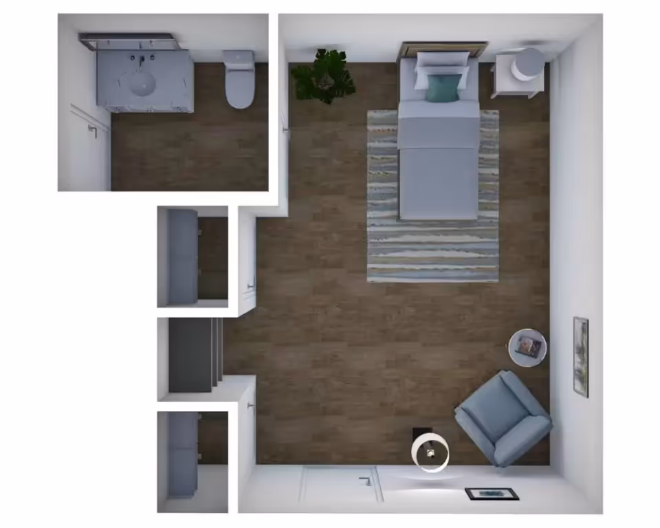 Top-down view of a small apartment floor plan featuring a bedroom area with a single bed, nightstand, armchair, and side table. Adjacent to the bedroom is a bathroom with a sink, mirror, and toilet. The floor is covered with wood-like flooring and there are a couple of framed pictures on the walls.