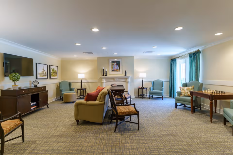 A spacious senior living common area with beige walls and carpeted floor. The room features a beige sofa with red pillows, several armchairs in light blue, a wooden TV stand with a flat-screen TV mounted above it, two side tables with lamps, a fireplace with decorative candles and a framed picture above it, and a wooden table with a chess set near a window with teal curtains.