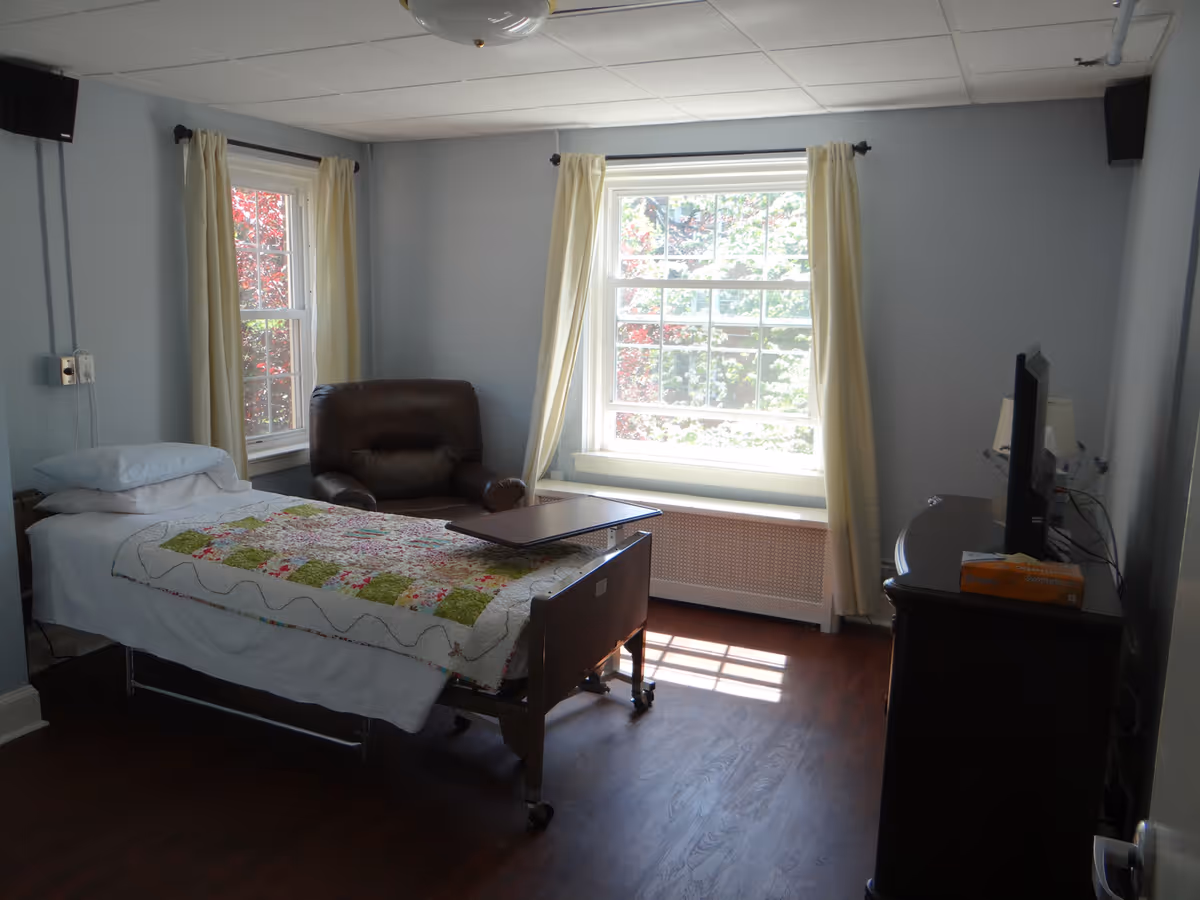 A hospice patient room with a single hospital bed covered with a colorful quilt, a brown leather armchair next to two windows with cream curtains, a wooden floor, and a dresser with a television on top.