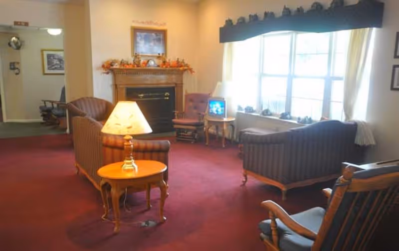 A cozy living room with a red carpet, two striped sofas, a wooden rocking chair, a small wooden side table with a lamp, a fireplace decorated with autumn-themed items, and a window with curtains letting in natural light.