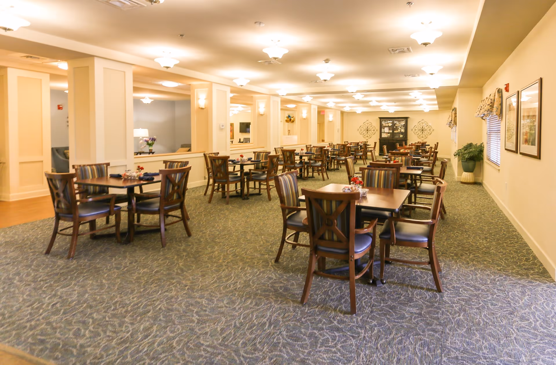 A spacious dining room with multiple wooden tables and chairs arranged neatly. The chairs have striped upholstery and the tables have small flower arrangements. The room is well-lit with ceiling lights and has carpeted flooring with a patterned design. There are windows with valances on the right wall and decorative wall art on the far wall.