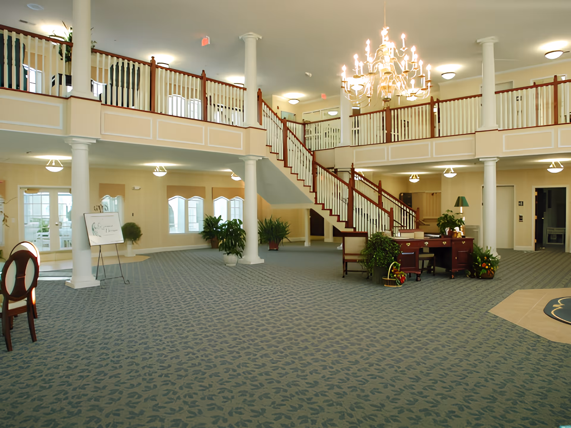 Spacious and well-lit interior of a senior living facility featuring a large open area with patterned carpet, white columns, a wooden staircase with white railings leading to an upper balcony, a chandelier hanging from the ceiling, several potted plants, and a wooden desk with chairs and decorative items.