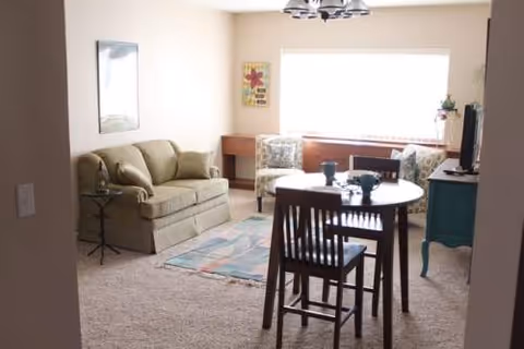 Sunlit living room with beige sofa, chairs, rug, and a round dining table.