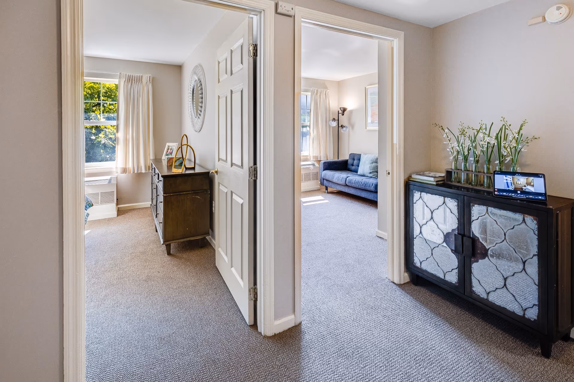 View of an interior hallway in a senior living facility with two open doorways. The left doorway leads to a bedroom with a dresser, window with curtains, and a round wall mirror. The right doorway leads to a living room area with a blue couch, floor lamp, and window with curtains. A decorative cabinet with glass doors and vases with flowers is positioned against the hallway wall, with a tablet placed on top displaying an image.