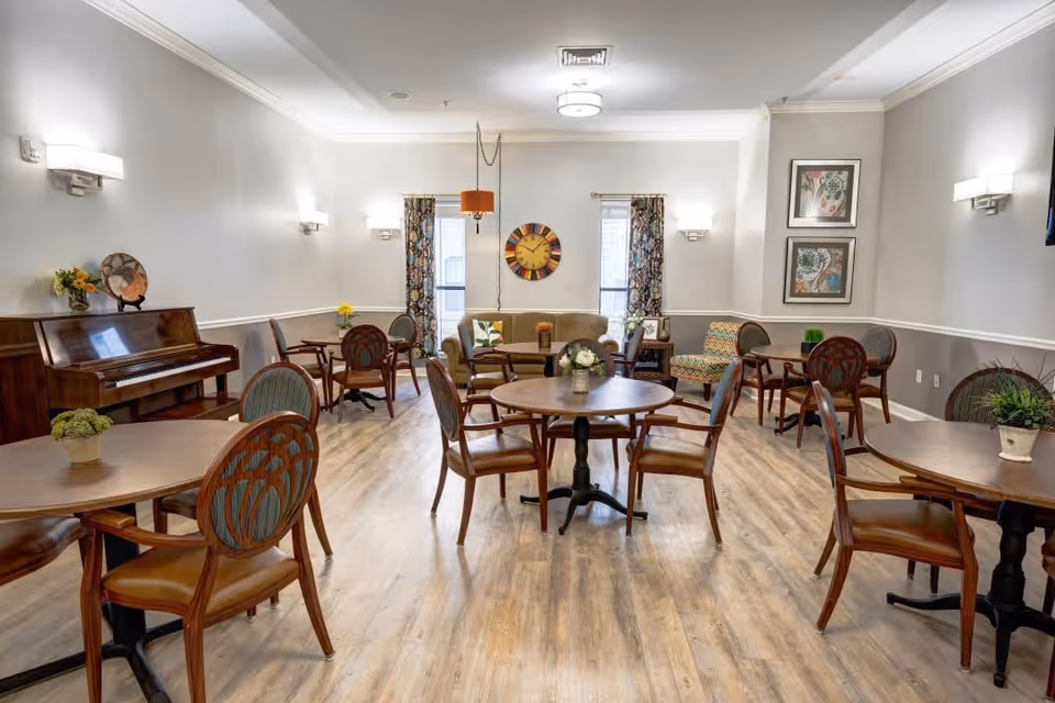 A bright and spacious common area with multiple round wooden tables and chairs arranged on a light wood floor. The room features a piano on the left side, a beige couch and patterned armchairs near two windows with floral curtains. The walls are painted light gray with white trim, decorated with framed artwork and a large colorful clock. Soft wall sconces and a ceiling light illuminate the room.