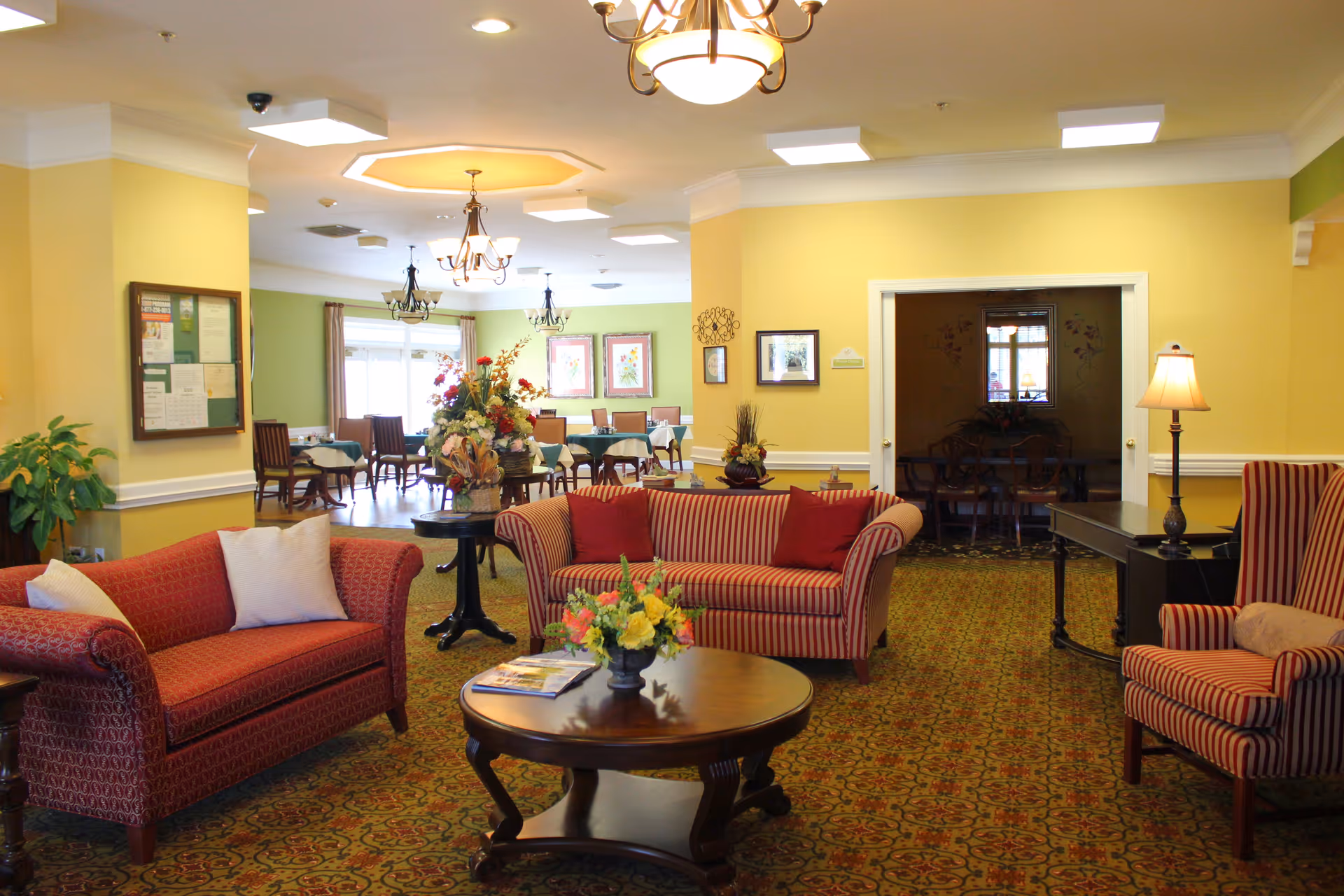 Bright senior living common lounge with upholstered sofas, armchairs, a coffee table and dining tables visible in the background.