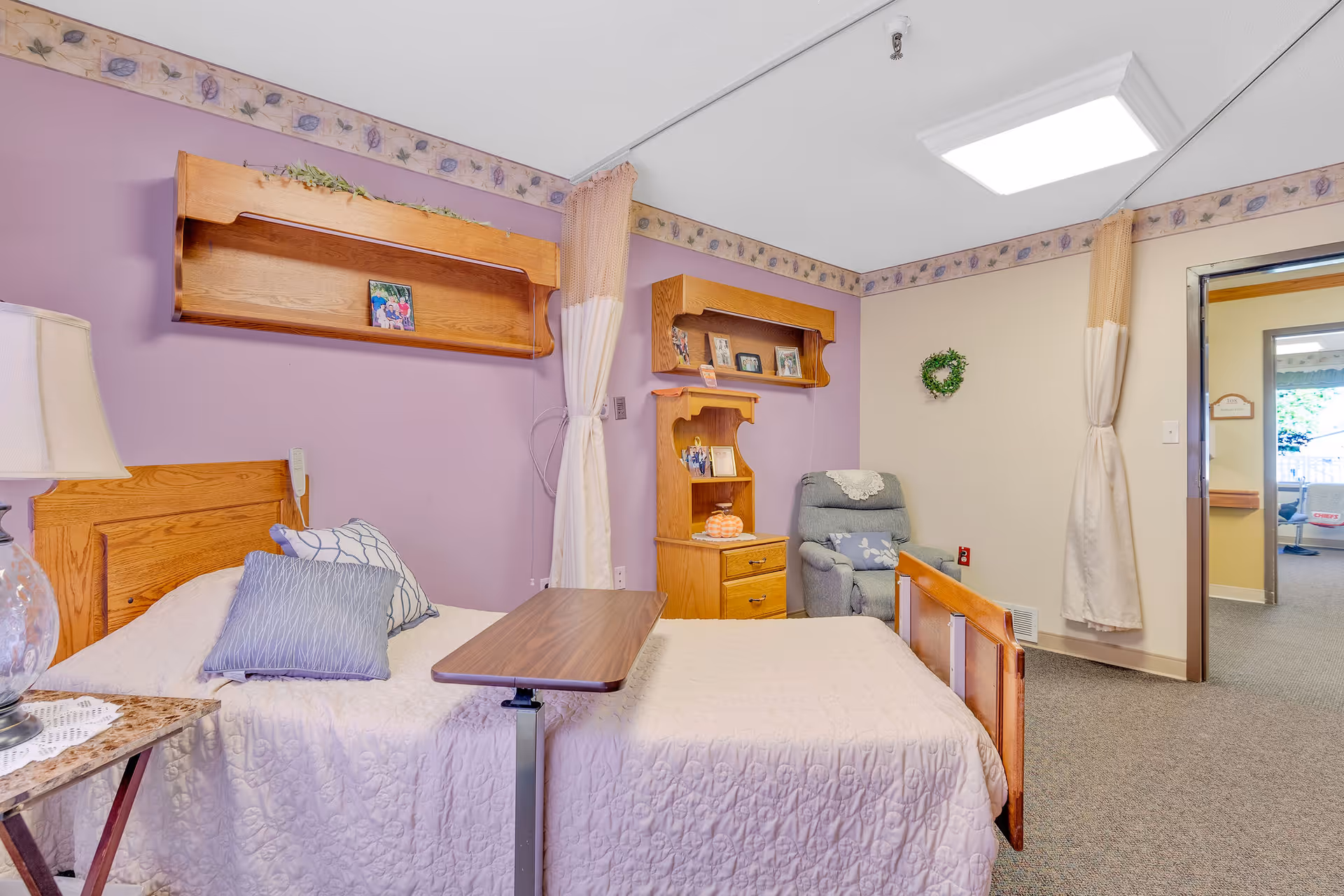 A cozy bedroom in a senior living facility with a single bed covered in a white quilt and two pillows. There is a wooden headboard and a small overbed table. The walls are painted purple and beige with floral wallpaper border near the ceiling. Wooden shelves with framed photos and decorative items are mounted on the walls. A comfortable gray recliner chair is placed in the corner near a window with beige curtains. The room has carpeted flooring and an open doorway leading to another area.