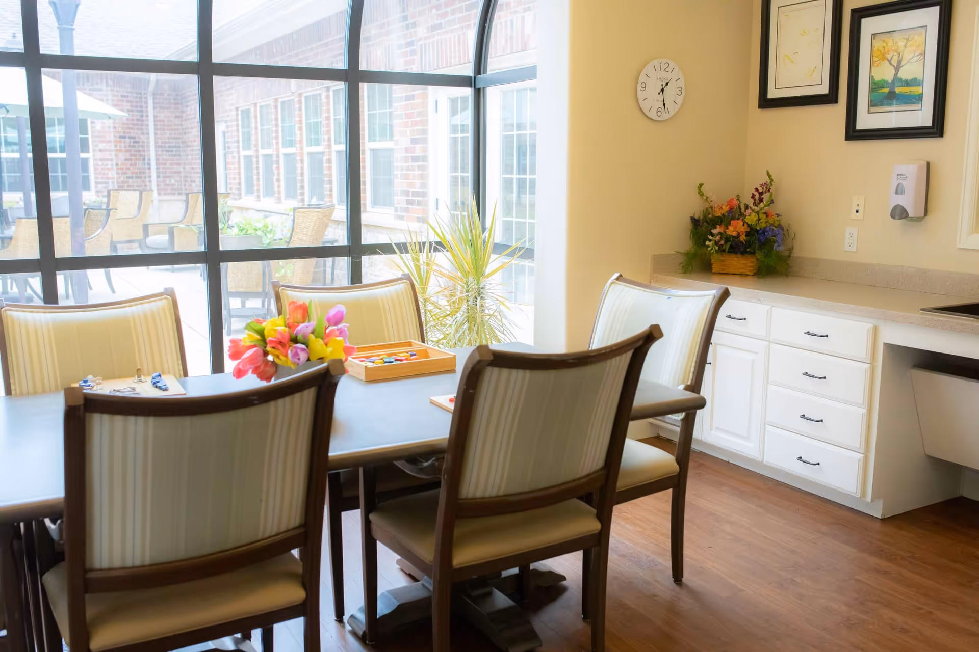 A bright dining area with a table and upholstered chairs, a vase of flowers on the table, countertop cabinets, and large windows overlooking a patio.