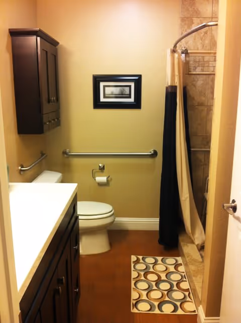 A small bathroom with beige walls and wooden floor. There is a white toilet with a metal grab bar behind it and a toilet paper holder on the wall. To the left is a dark wooden vanity with a white countertop and a matching wall cabinet above it. On the right side, there is a shower area with beige tile walls and a curved shower rod holding a beige and black shower curtain. A small rug with a circular pattern is placed on the floor in front of the shower. A framed picture hangs on the wall above the toilet.