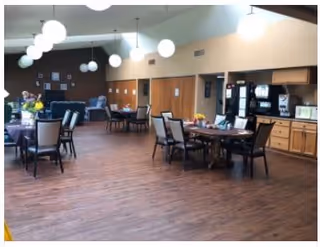 A spacious senior living common area with wooden flooring, multiple tables and chairs arranged for dining or socializing, a beverage station with coffee machines and a microwave on the right side, and modern spherical pendant lights hanging from the ceiling.