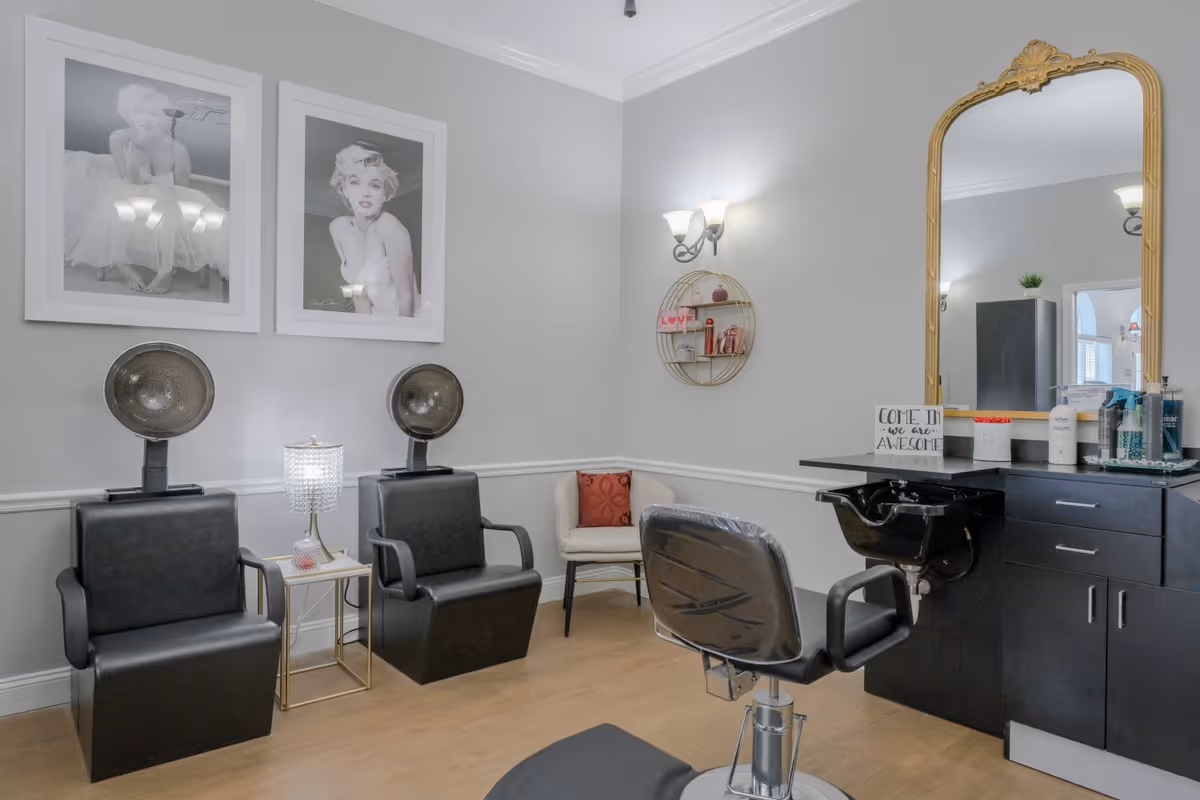A salon room with two black hooded hair dryers, a styling chair and wash sink, a large gold-framed mirror and black-and-white portrait prints on the wall.