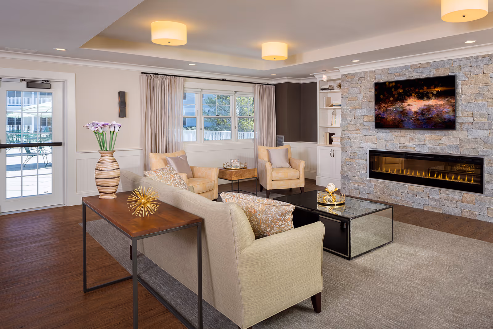 Bright communal living room with sofas and armchairs, a glass coffee table, and a stone wall fireplace with a mounted TV.