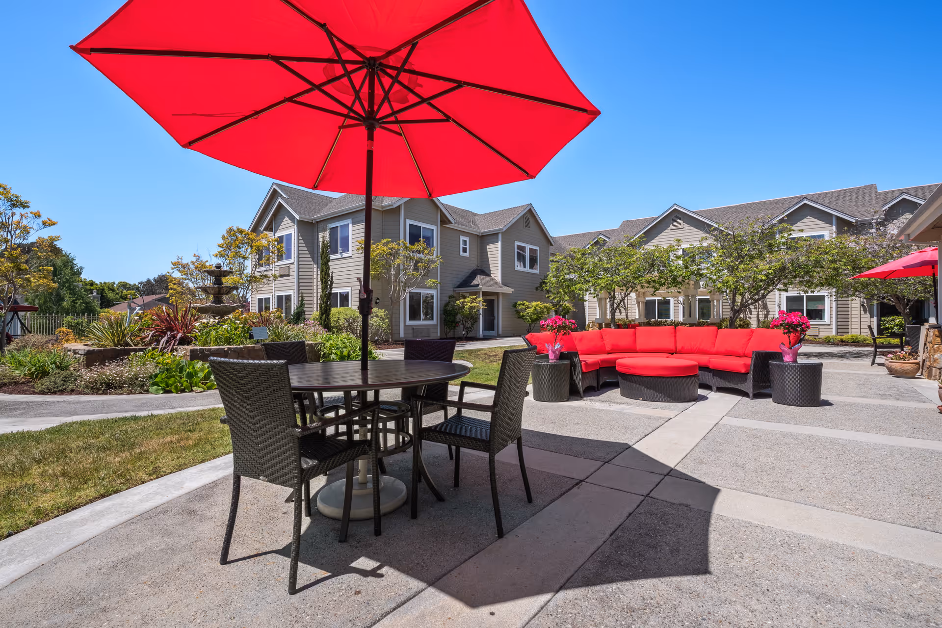 Outdoor courtyard with red umbrellas, a round table and chairs, and a red sectional sofa in front of multi-story residential buildings.