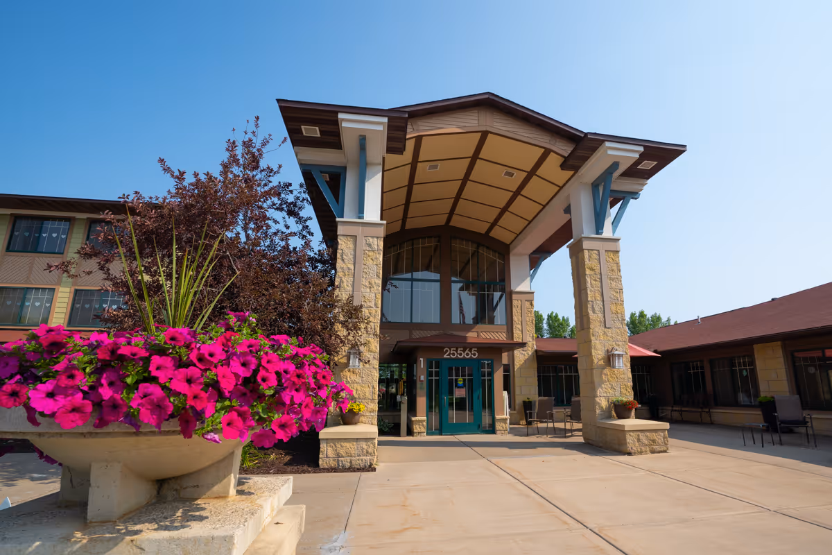 Entrance of a senior living facility with a covered porch supported by stone pillars. There is a large planter with vibrant pink flowers in the foreground and the building number 25565 above the glass doors. The sky is clear and blue.