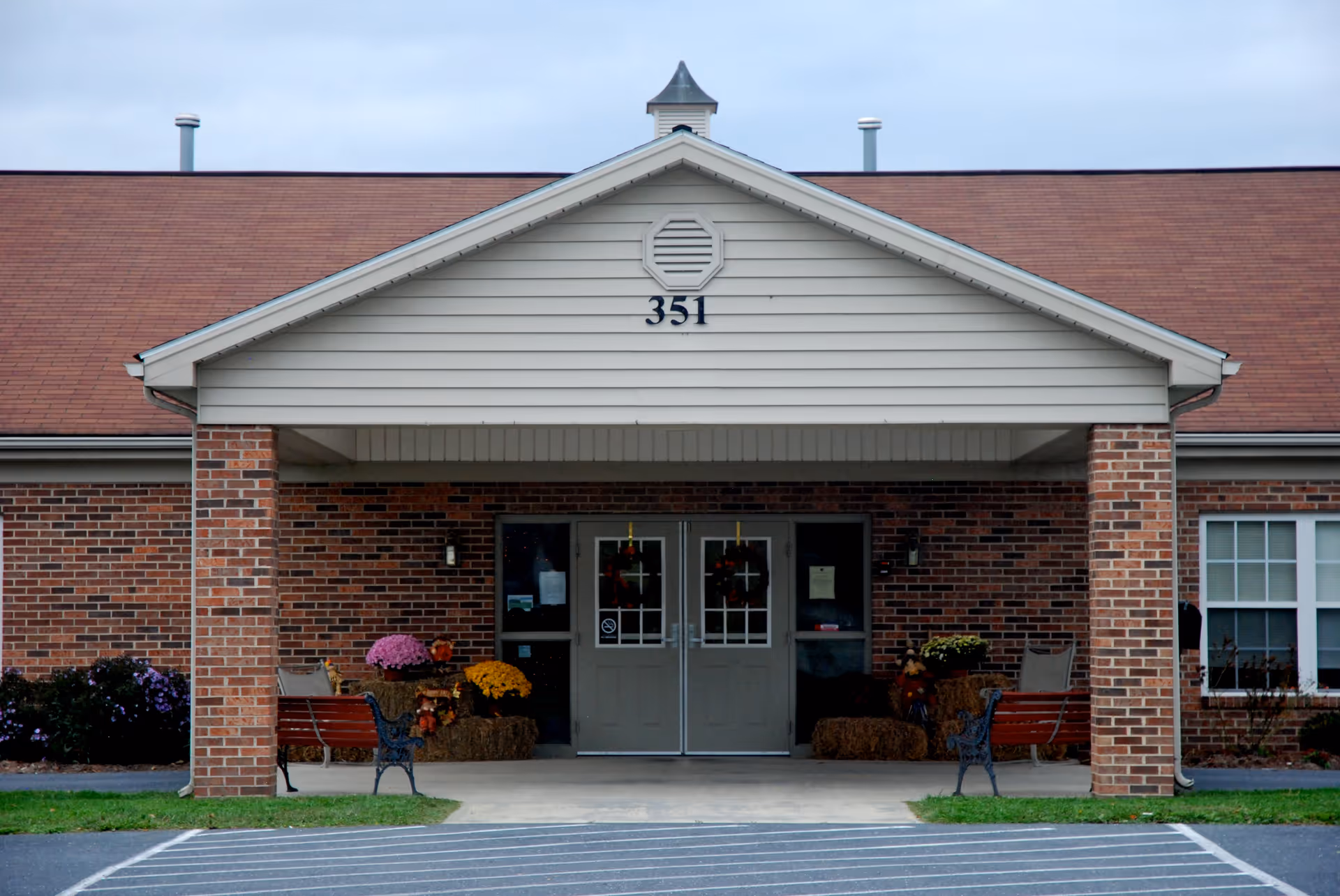 Front entrance of a brick building with the number 351 displayed above the double doors. The entrance is covered by a white gabled roof supported by brick columns. There are benches and autumn-themed decorations including flowers and hay bales on either side of the entrance.
