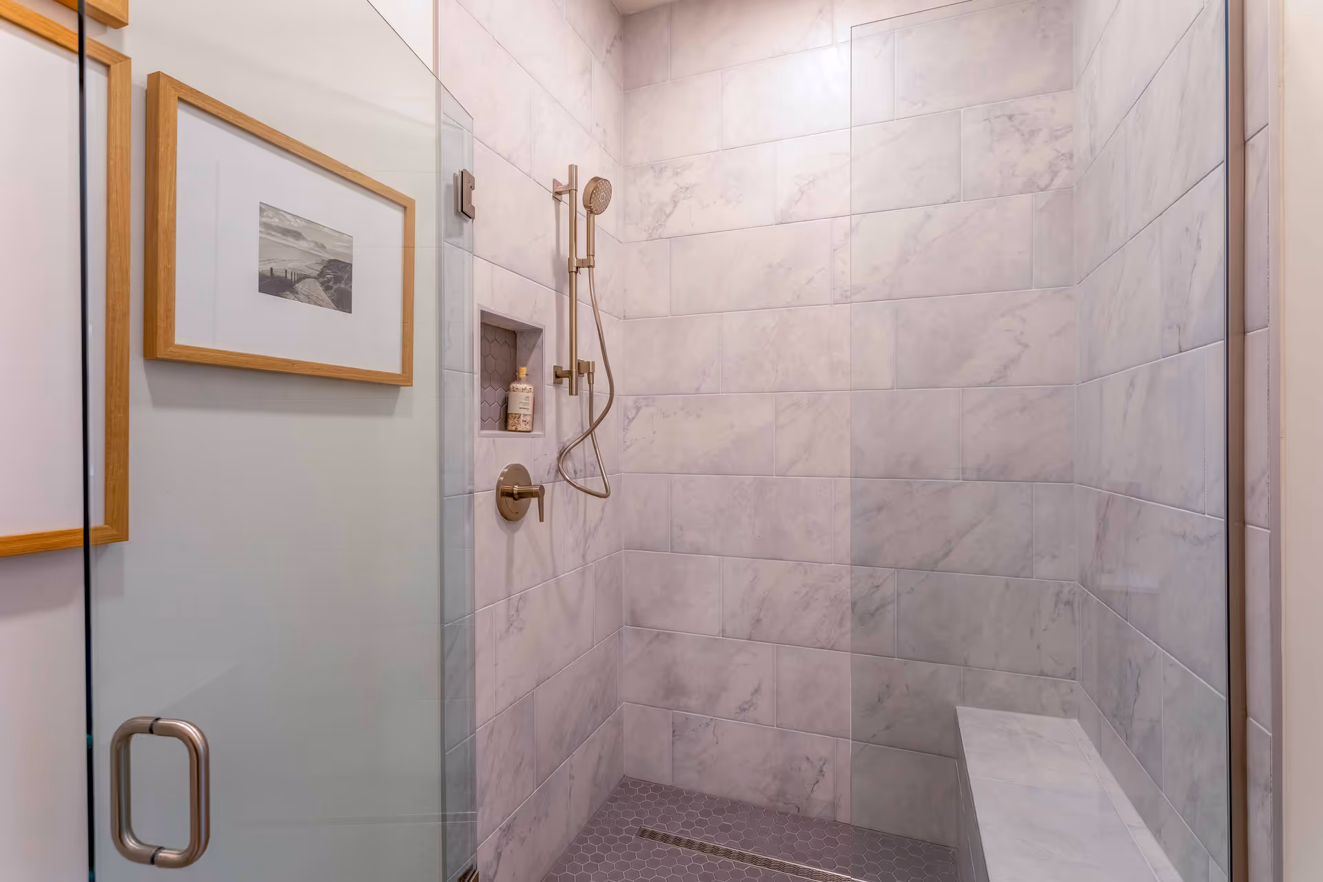 A modern shower area with large rectangular marble tiles on the walls and small hexagonal tiles on the floor. The shower features a handheld showerhead with a metal hose and a built-in niche holding a bottle. There is a glass door with a metal handle and a bench on the right side. Two framed pictures with wooden frames hang on the wall outside the shower.