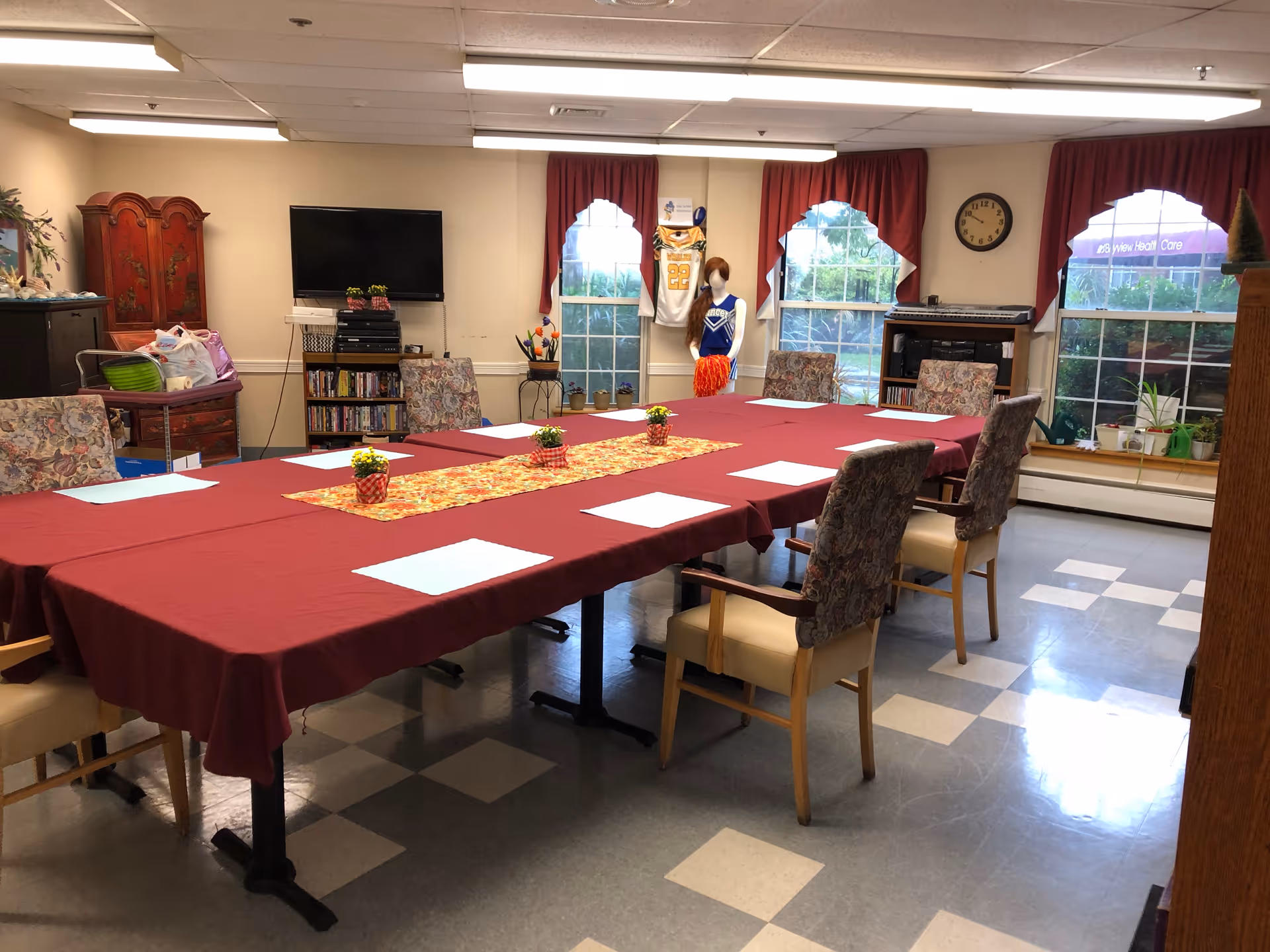 A meeting or activity room with a large rectangular table covered with a red tablecloth and a decorative runner. Several chairs with floral upholstery surround the table. The room has three windows with red curtains, a wall clock, a TV mounted on the wall, a bookshelf with DVDs, and a mannequin dressed in a cheerleader outfit. There are small potted plants on the table and on the windowsill.