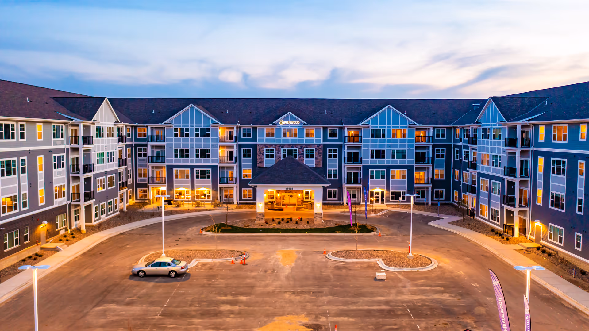 Exterior view of Havenwood of Maple Grove senior living facility at dusk, showing a large multi-story building with many windows lit from inside, a covered entrance, and an empty parking lot in front.