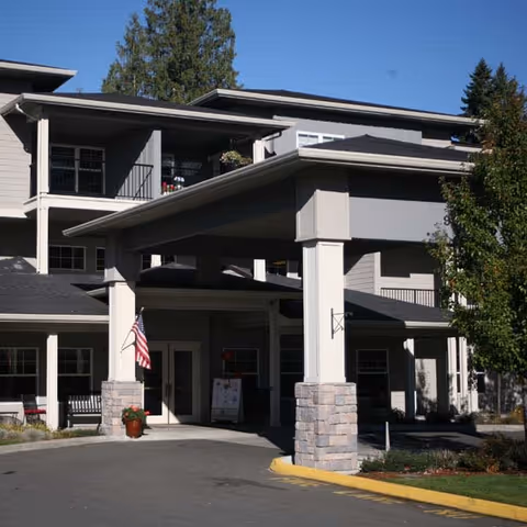 Exterior view of a multi-story senior living facility with a covered entrance supported by large pillars. The building is light gray with stone accents at the base of the pillars. An American flag is displayed near the entrance, and there are trees and landscaping around the driveway.