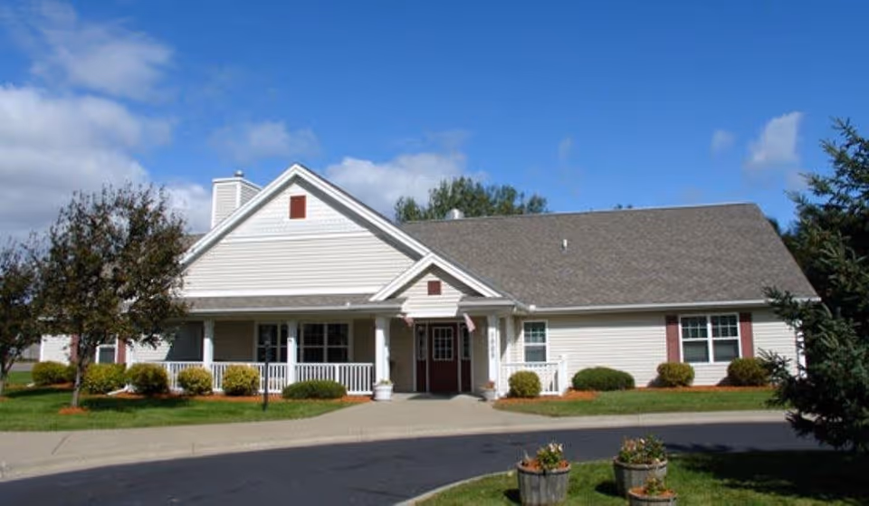 Exterior view of a single-story residential building with white siding, a gray shingled roof, and a covered front porch with white railings. The building is surrounded by green grass, small bushes, and trees under a partly cloudy blue sky.