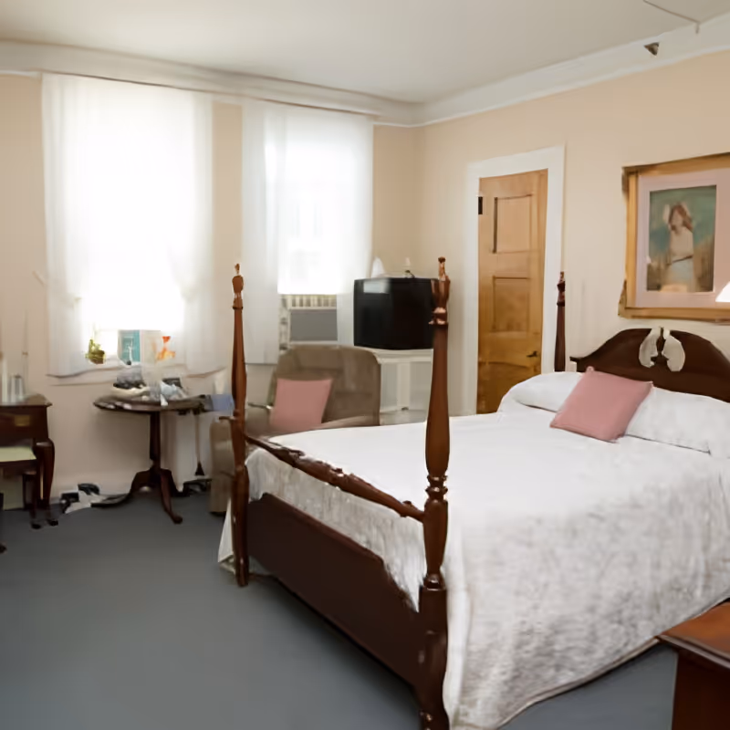 Sunlit bedroom with a wooden four-poster bed dressed in a white coverlet and pink pillows, an armchair and small table, and a TV under curtained windows.