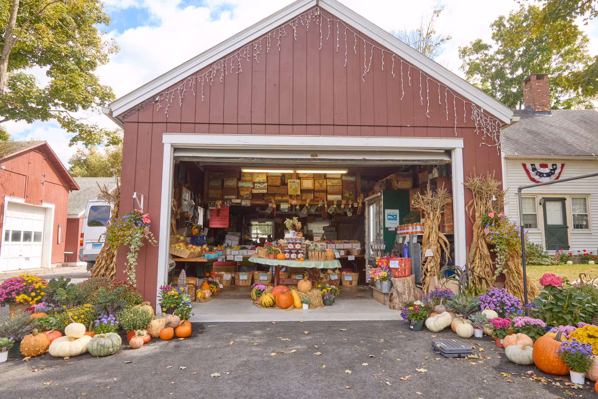 Open red barn-style building with a variety of pumpkins, gourds, flowers, and plants arranged outside and inside. The interior has tables with jars and baskets of produce, and the exterior is decorated with hanging plants and cornstalks. The scene is bright and sunny with trees and a white house visible in the background.
