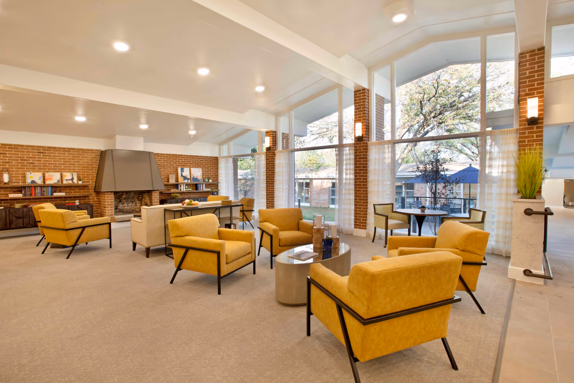 A spacious and well-lit living room area with large floor-to-ceiling windows showing an outdoor view with trees and umbrellas. The room features yellow armchairs arranged around a small round coffee table, a fireplace with a modern hood, bookshelves with books and decorative items, and additional seating with tables and chairs near the windows.