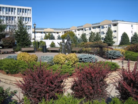 Outdoor garden area with various shrubs and bushes, a paved walkway, benches, and a statue of two figures in the center. In the background, there are multi-story residential buildings under a clear blue sky.