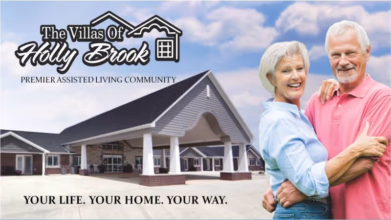 An elderly couple smiling in front of the Villas of Holly Brook assisted living building and logo.