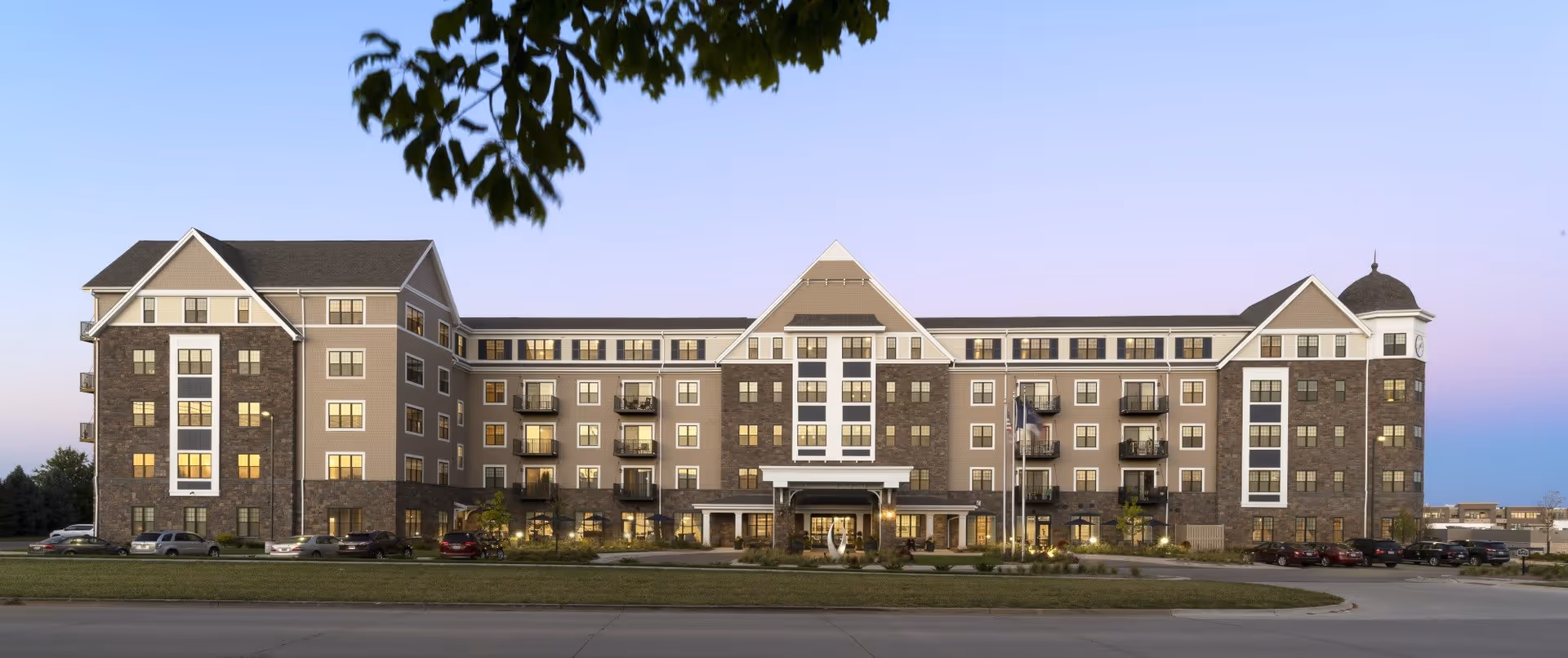 Front exterior view of a large, multi-story senior living facility building named Grand Living At Tower Place, with a mix of stone and beige siding, multiple windows, balconies, and a clock tower on the right side. Several cars are parked in front, and there is a tree branch partially visible at the top.