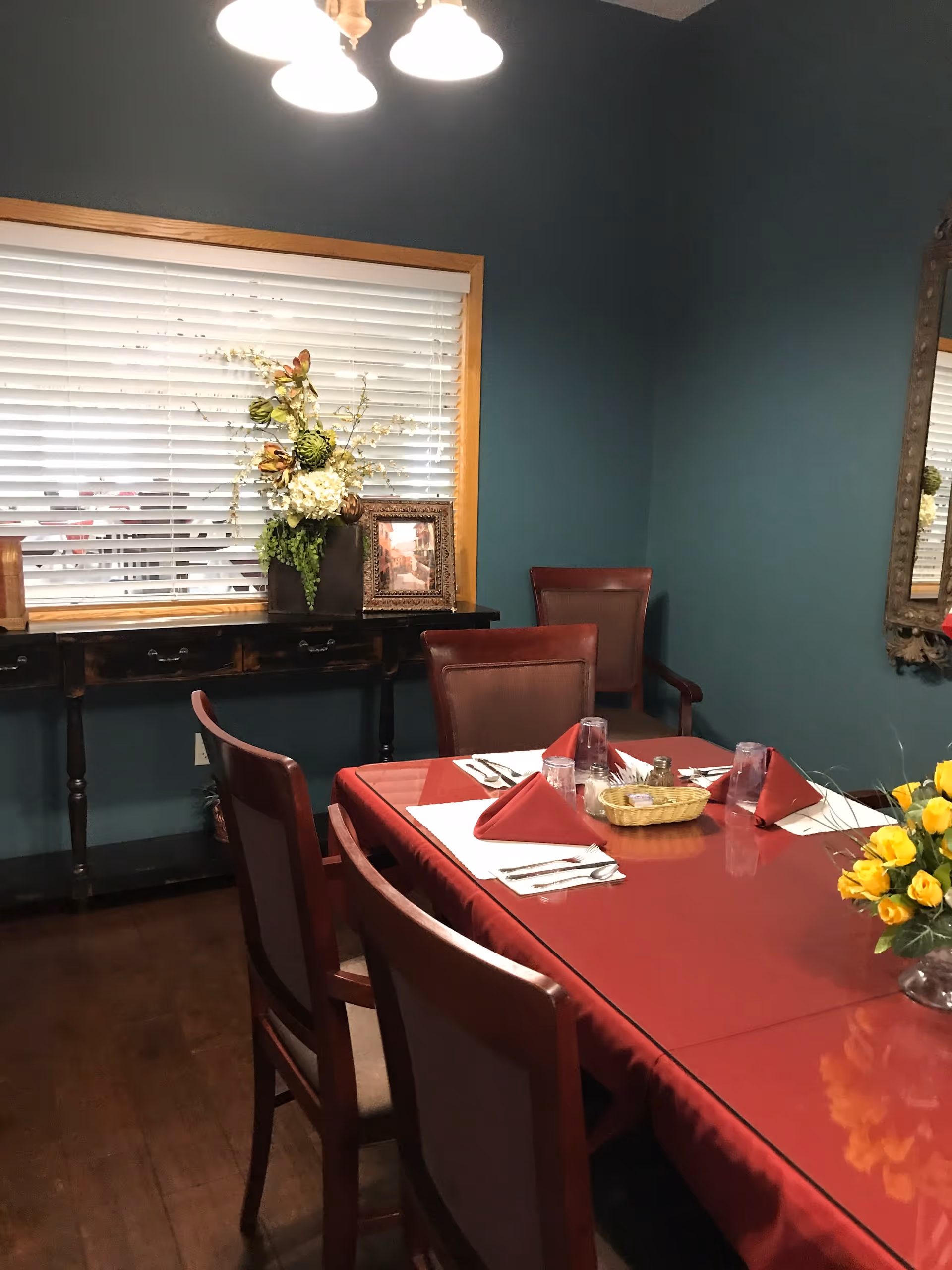 A dining room with a rectangular table covered with a red tablecloth, set with red folded napkins, glasses, and silverware. There are six wooden chairs with cushioned seats around the table. A side table against a teal wall holds a floral arrangement and a framed picture. A window with white blinds is above the side table, and a decorative mirror hangs on the adjacent wall. A ceiling light fixture with three bulbs illuminates the room.