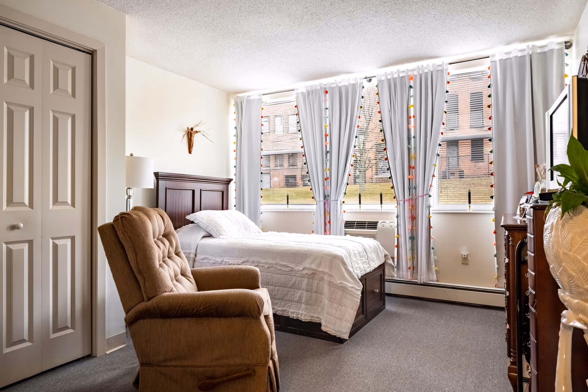 A cozy senior living bedroom with a single bed covered in white bedding, a cushioned recliner chair, a wooden dresser with a large mirror, and large windows with white curtains decorated with colorful string lights. The room has neutral walls and carpeted flooring.