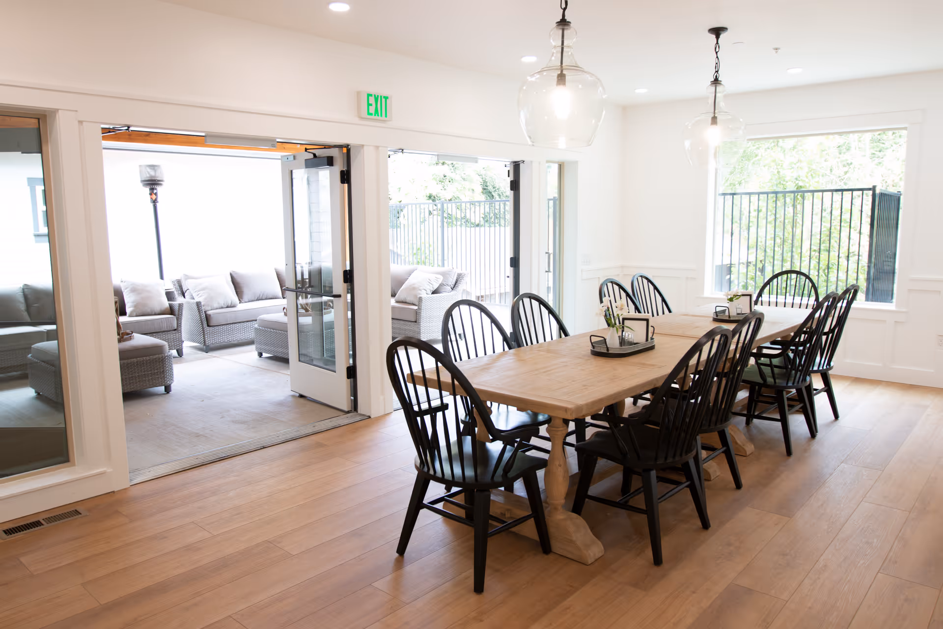 A bright dining room with a long wooden table surrounded by black chairs. Two large glass pendant lights hang from the ceiling. The room has light wood flooring and white walls with a large window on the right. Double glass doors open to an outdoor patio area furnished with cushioned wicker seating and a tall outdoor heater.