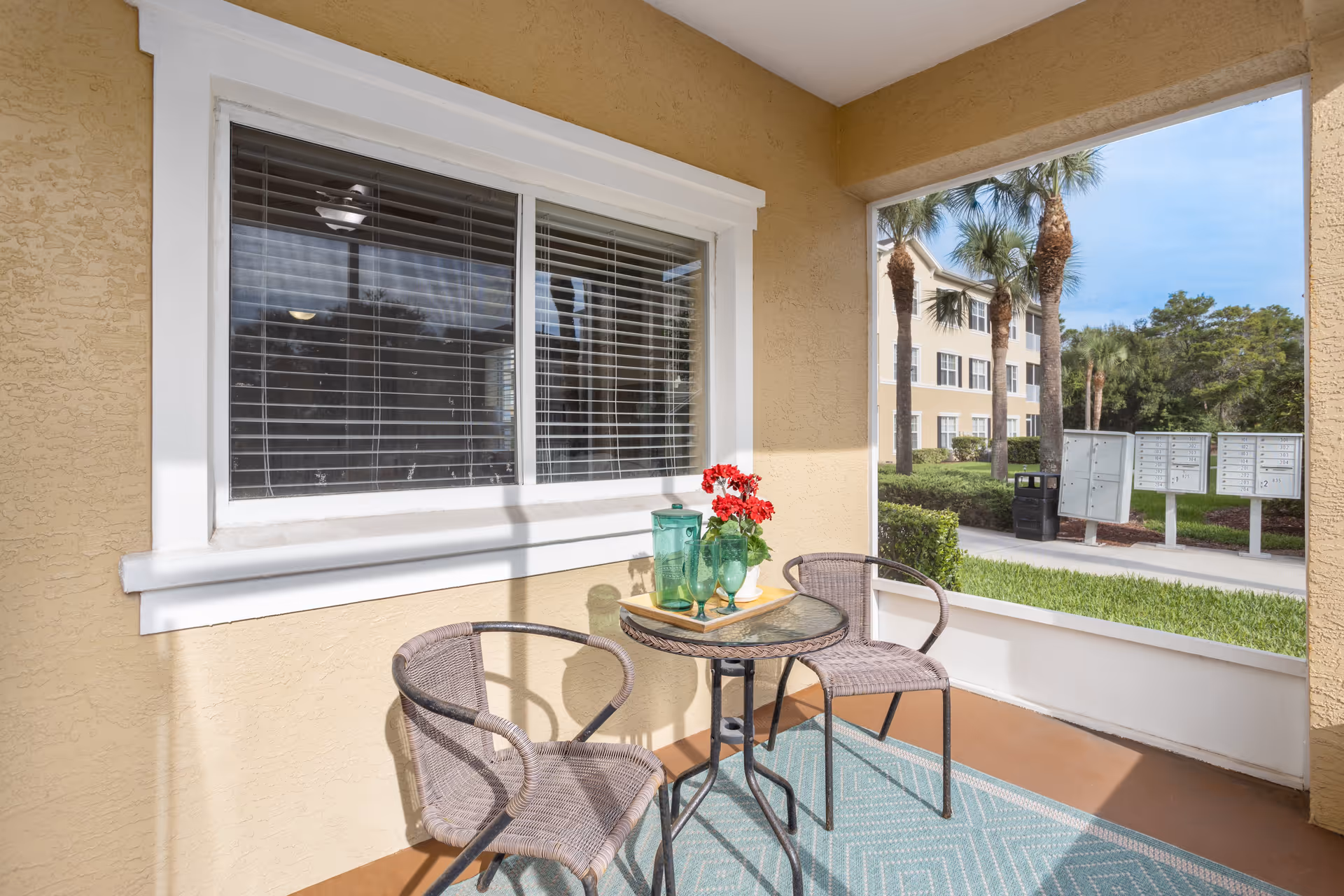 A small outdoor patio area with two wicker chairs and a round glass-top table. On the table, there are green glassware and a small vase with red flowers. The patio has a beige textured wall with a white-framed window with blinds. In the background, there are palm trees, a grassy area, and a set of mailboxes near a sidewalk.