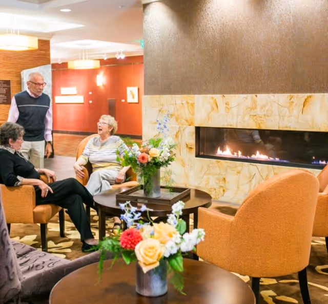 Three elderly people sitting and standing around a round table with floral arrangements in a cozy living room area with a modern fireplace and warm lighting.