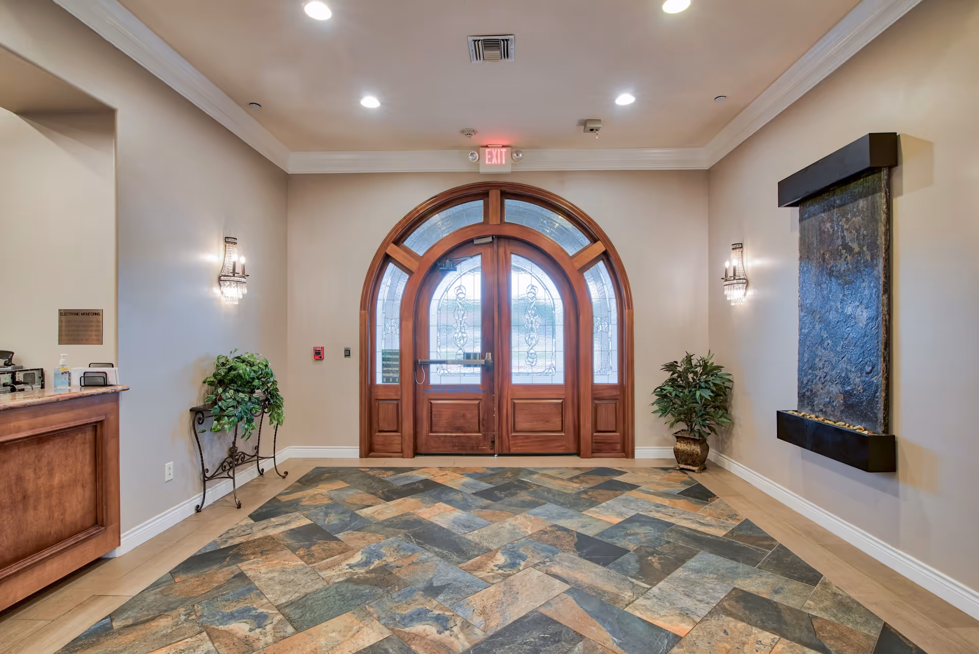 Entrance area of Pelican Bay Assisted Living Community featuring a large wooden double door with decorative glass panels, a tiled floor with a mix of earth tones, a water feature mounted on the right wall, two wall sconces with lights, a small reception desk on the left, and potted plants on either side of the door.