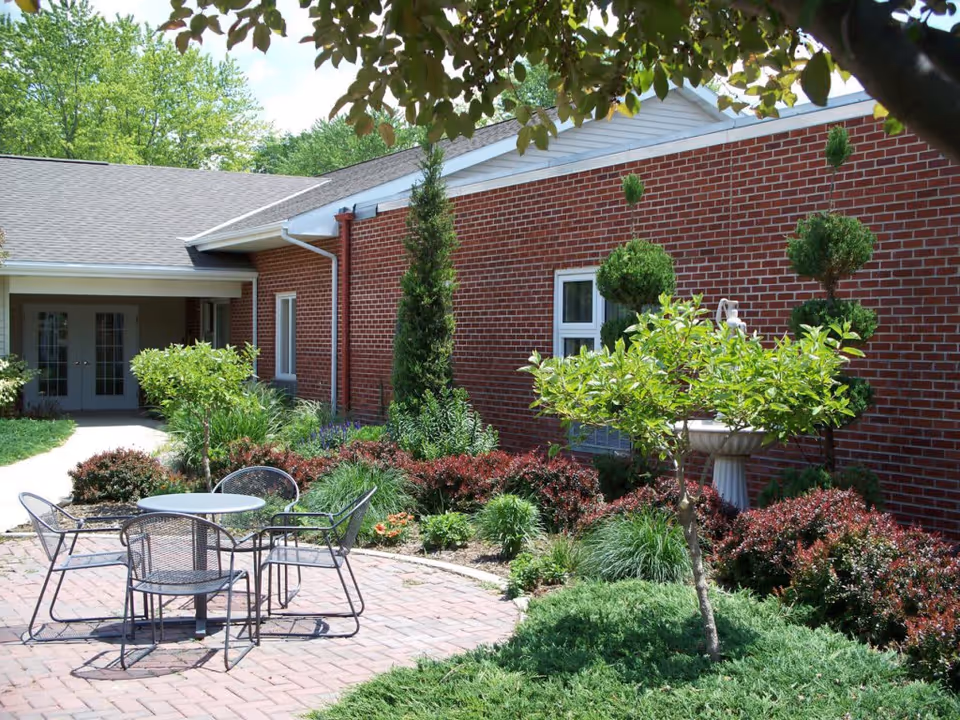 Outdoor patio area at Regency Square with a round metal table and four matching chairs on a brick-paved surface, surrounded by well-maintained shrubs, small trees, and a brick building with windows in the background.