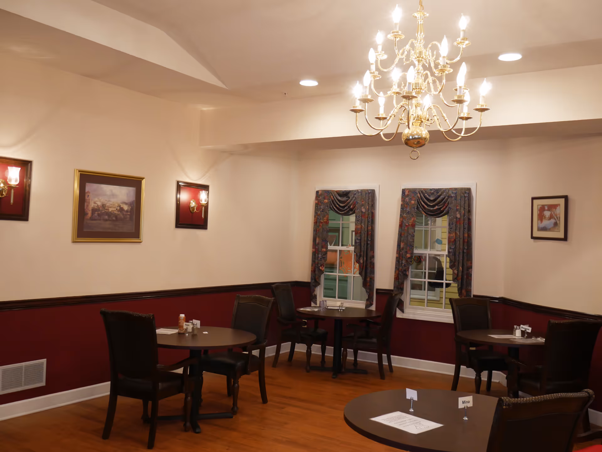 A dining room with several round tables and dark wooden chairs. The walls are painted beige with a dark red wainscoting. There are framed pictures and wall sconces on the walls. Two windows with floral curtains are visible, and a large chandelier hangs from the ceiling. The floor is wooden.