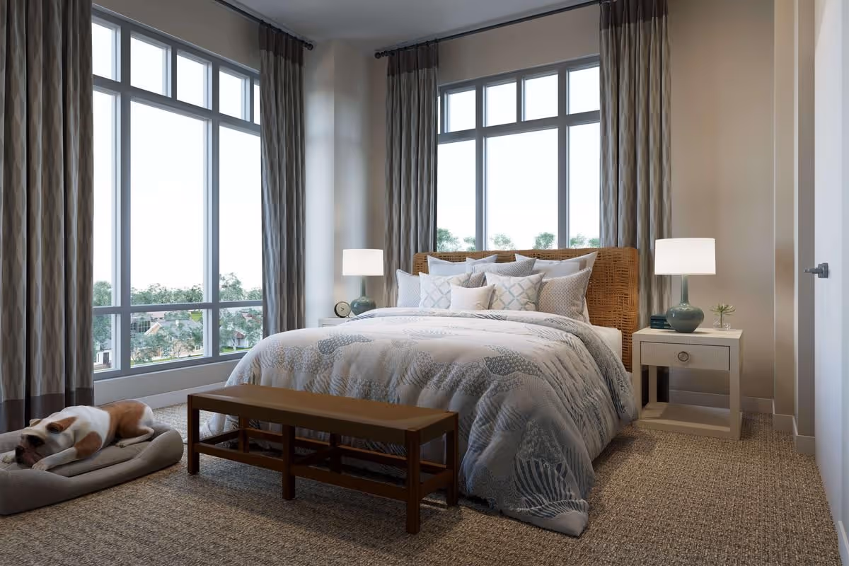 A cozy bedroom with a large bed covered in patterned bedding and multiple pillows. There are two bedside tables with lamps on each side of the bed. A wooden bench is placed at the foot of the bed. Large windows with curtains allow natural light to fill the room. A dog is resting on a pet bed on the carpeted floor near the window.