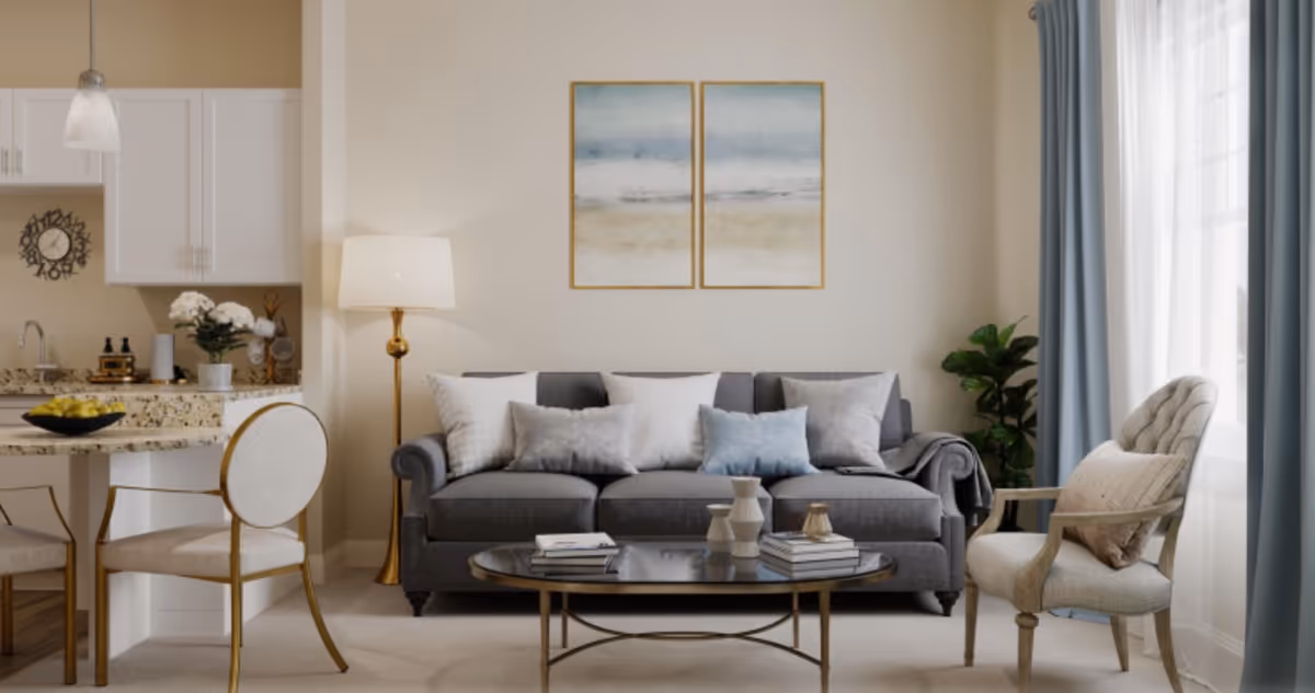 Bright, neutrally decorated living room with a gray sofa, coffee table, accent chairs, floor lamp, and a small kitchen counter visible to the left.