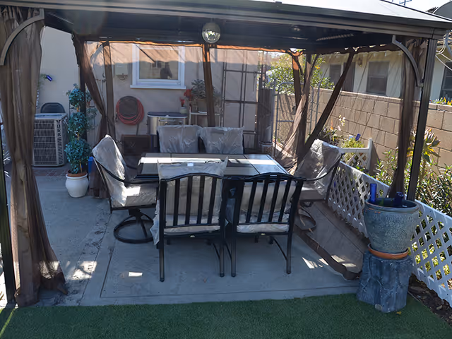 Outdoor patio area with a covered gazebo featuring a glass-top table and six cushioned chairs. The patio is surrounded by a white lattice fence and potted plants, with a concrete floor and artificial grass in the foreground.