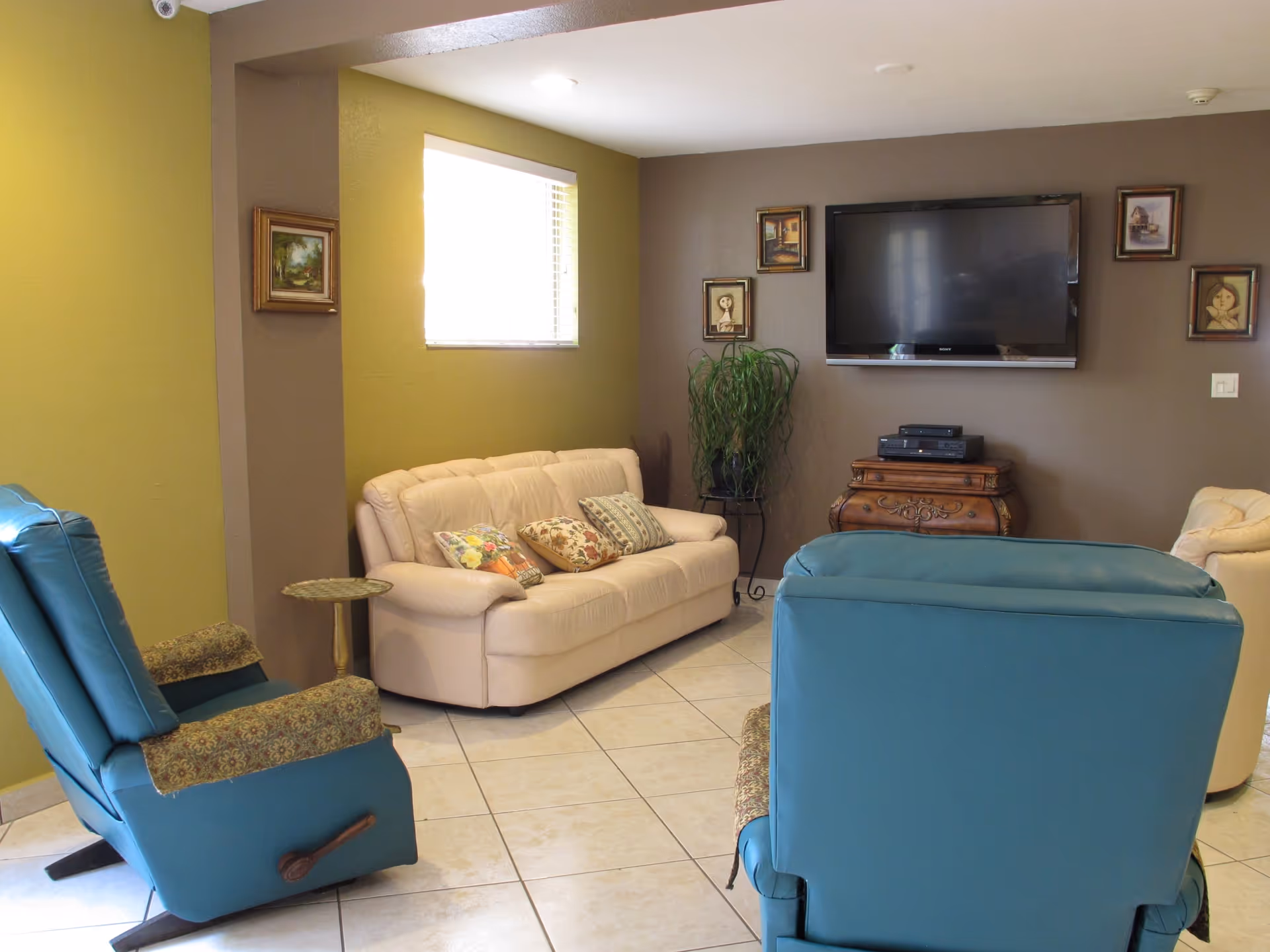 A cozy living room with a beige sofa adorned with floral and striped cushions, two blue recliner chairs with patterned armrest covers, a small round side table, a flat-screen TV mounted on a brown accent wall, and several framed paintings surrounding the TV. A potted plant is placed on a stand beneath the TV, and the room has tiled flooring and a window with blinds letting in natural light.