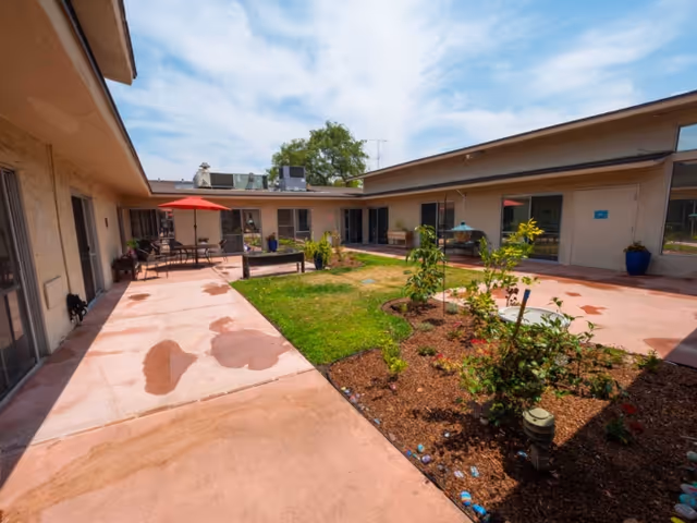 Outdoor courtyard area of a senior living facility with a pink concrete walkway surrounding a small garden with plants and grass. There are tables with umbrellas and chairs along the walkway, and the building forms a U-shape around the courtyard under a partly cloudy sky.