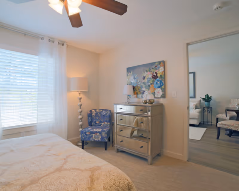 A bright bedroom with a bed in the foreground, a mirrored dresser, a floral armchair and floor lamp by a window, and a doorway leading to a sitting area.
