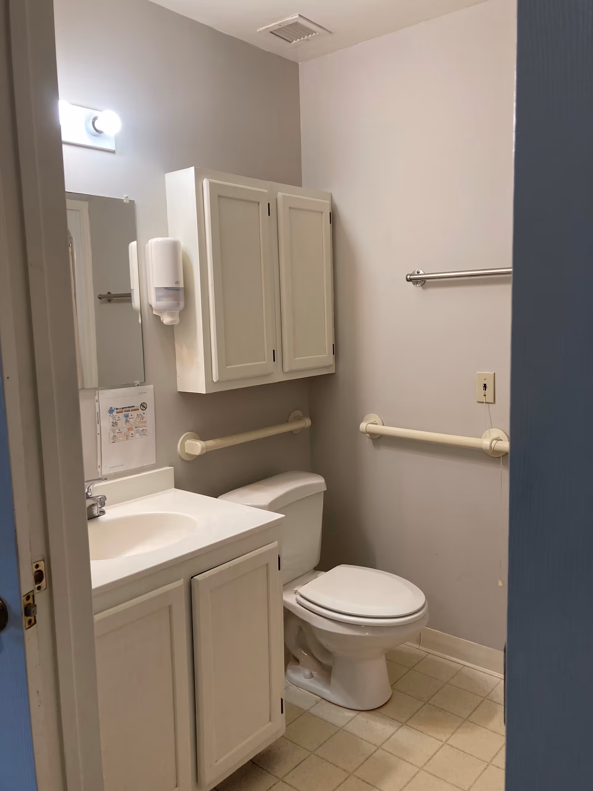 Small accessible bathroom with a toilet, sink vanity, wall-mounted cabinet, mirror, and grab bars.