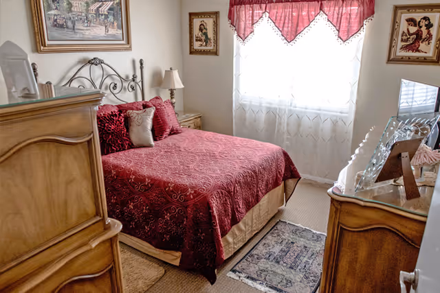 A cozy bedroom with a red bedspread and decorative pillows, wooden dressers, and a window with a red valance.